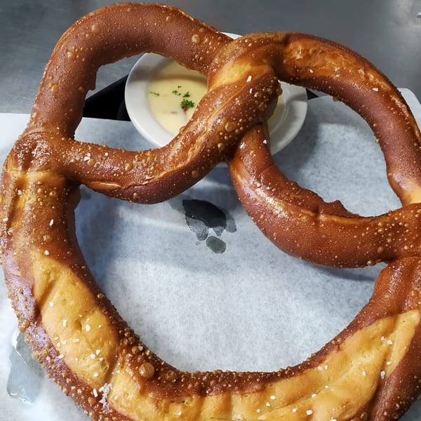 A large, salted pretzel with a side of creamy cheese dip garnished with herbs.