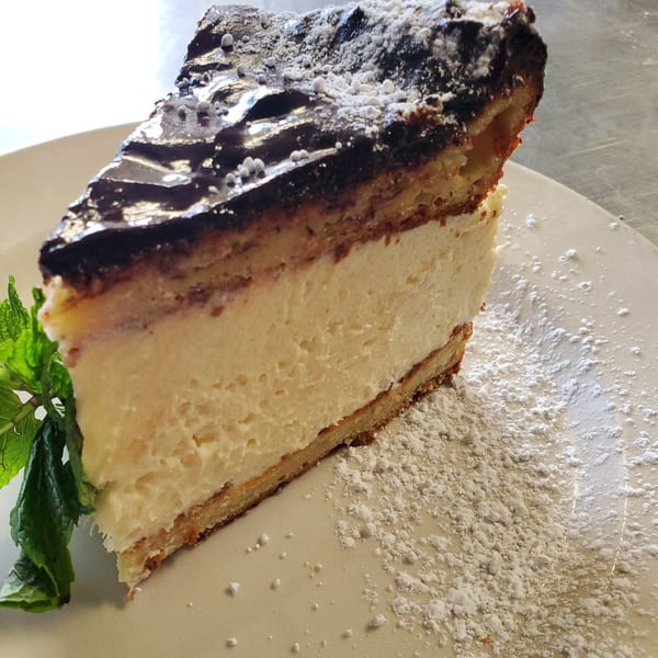 Slice of cheesecake with chocolate topping, powdered sugar, and a mint sprig on a white plate.