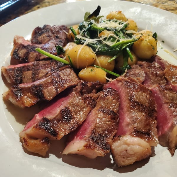 Sliced grilled steak with gnocchi, spinach, and grated cheese on a white plate.