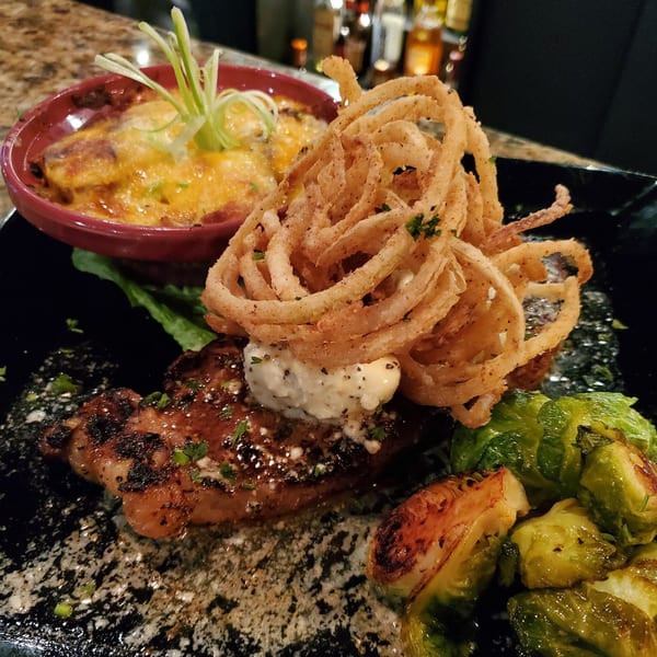 Southern-style steak with onion rings, cheesy grits, and roasted Brussels sprouts on a black plate.