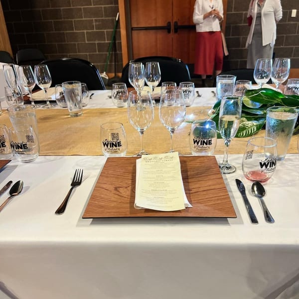 A formal table setting with wine glasses, water glasses, silverware, and a menu on a wooden charger.