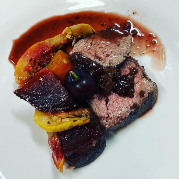 Seared beef slices with dark sauce, roasted vegetables, and a grape on a white plate.