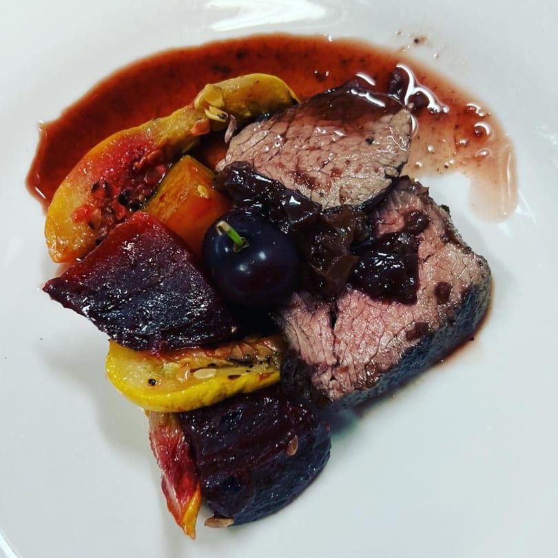 Seared beef slices with dark sauce, roasted vegetables, and a grape on a white plate.