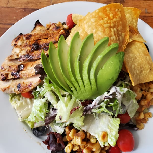 Grilled chicken salad with avocado, corn, tomatoes, and tortilla chips on a white plate.