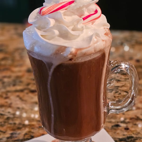 Hot chocolate with whipped cream and two candy canes in a clear mug on a speckled counter.