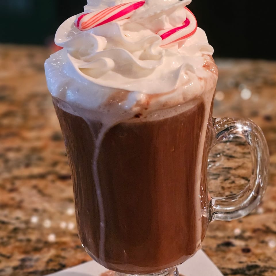 Hot chocolate with whipped cream and two candy canes in a clear mug on a speckled counter.