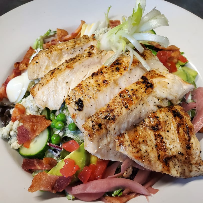 Grilled chicken salad with bacon, avocado, tomatoes, peas, blue cheese, and pickled onions on a white plate.
