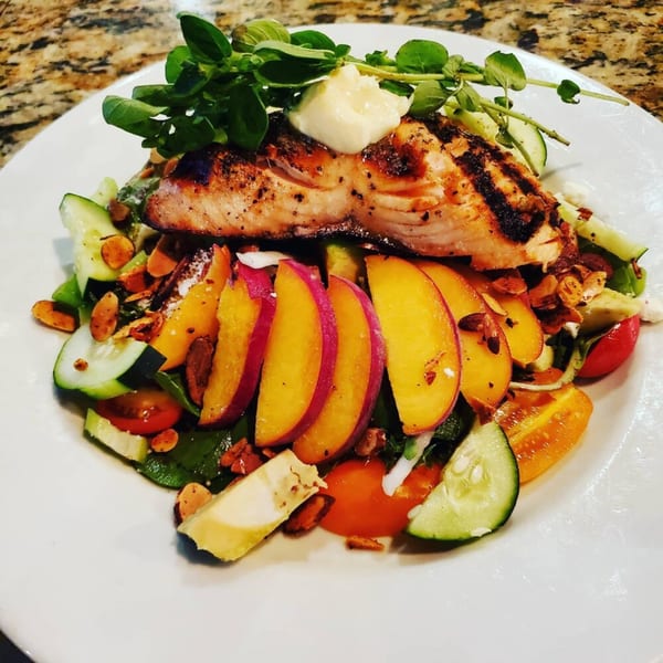 Grilled salmon with a dollop of sauce, fresh greens, sliced peaches, cucumbers, and tomatoes.