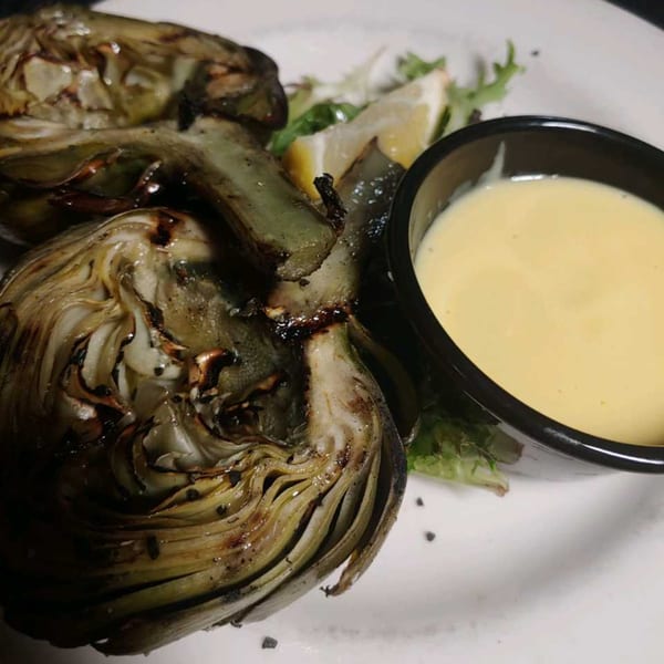 Grilled artichokes with a side of creamy dipping sauce, lemon wedges, and greens on a white plate.