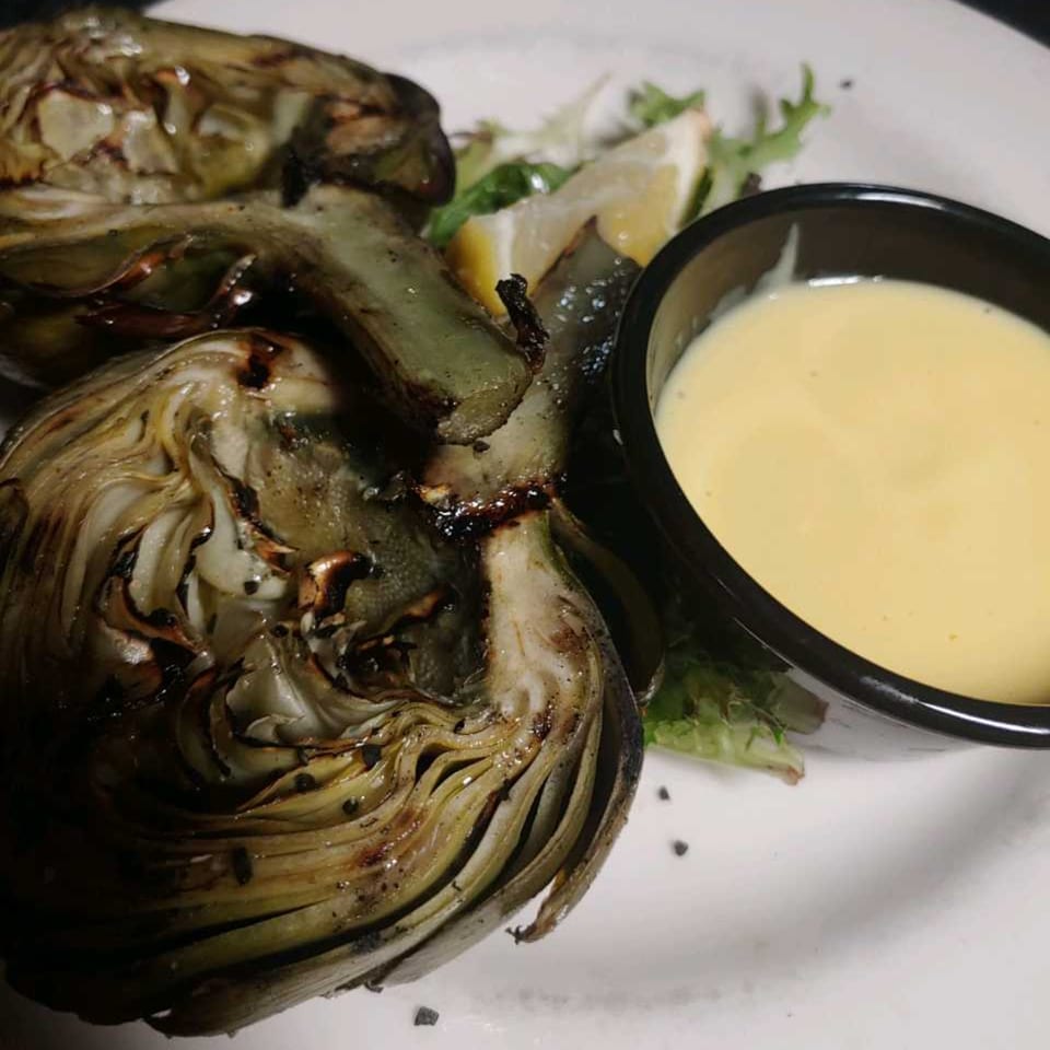 Grilled artichokes with a side of creamy dipping sauce, lemon wedges, and greens on a white plate.