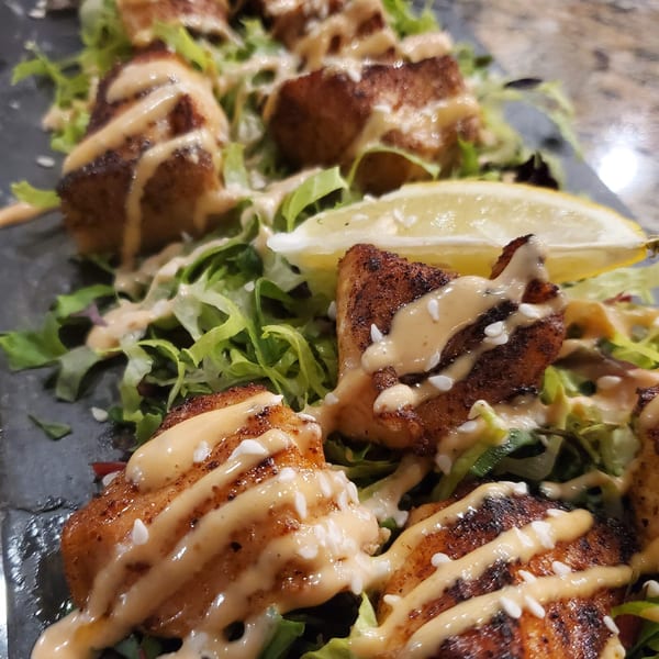 Southern-style blackened fish bites with creamy sauce, sesame seeds, and lettuce, garnished with a lemon wedge.